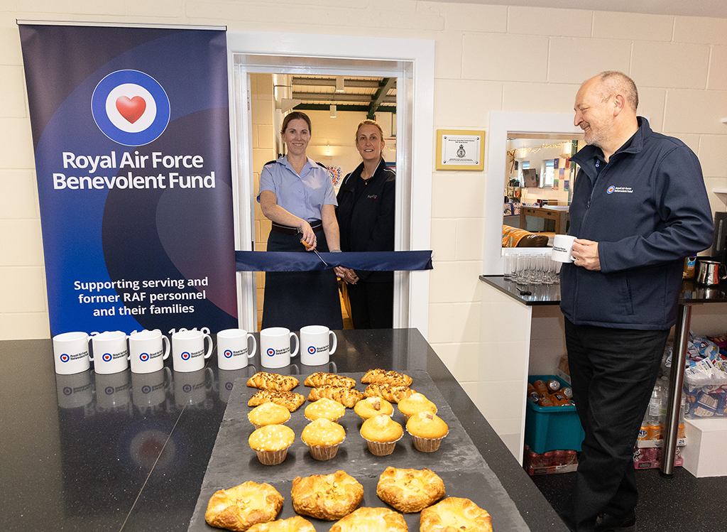 Our Station Commander cuts a ceremonial ribbon at the door of the MAC kitchen. A Serco and RAFBF representative stand with her. Cakes, pasties mugs and other fare are displayed in the foreground.
